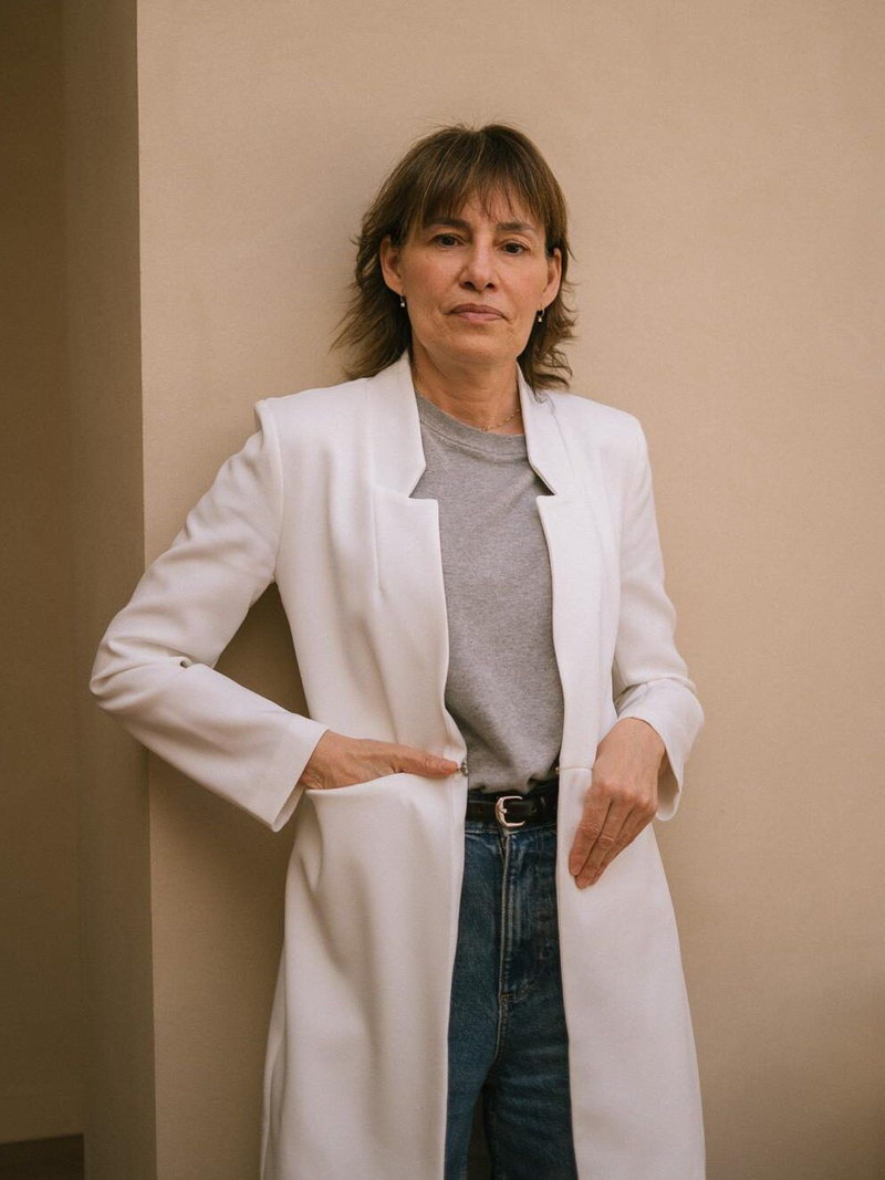 Montse Carreras de pie junto a una pared clara, con americana blanca larga.