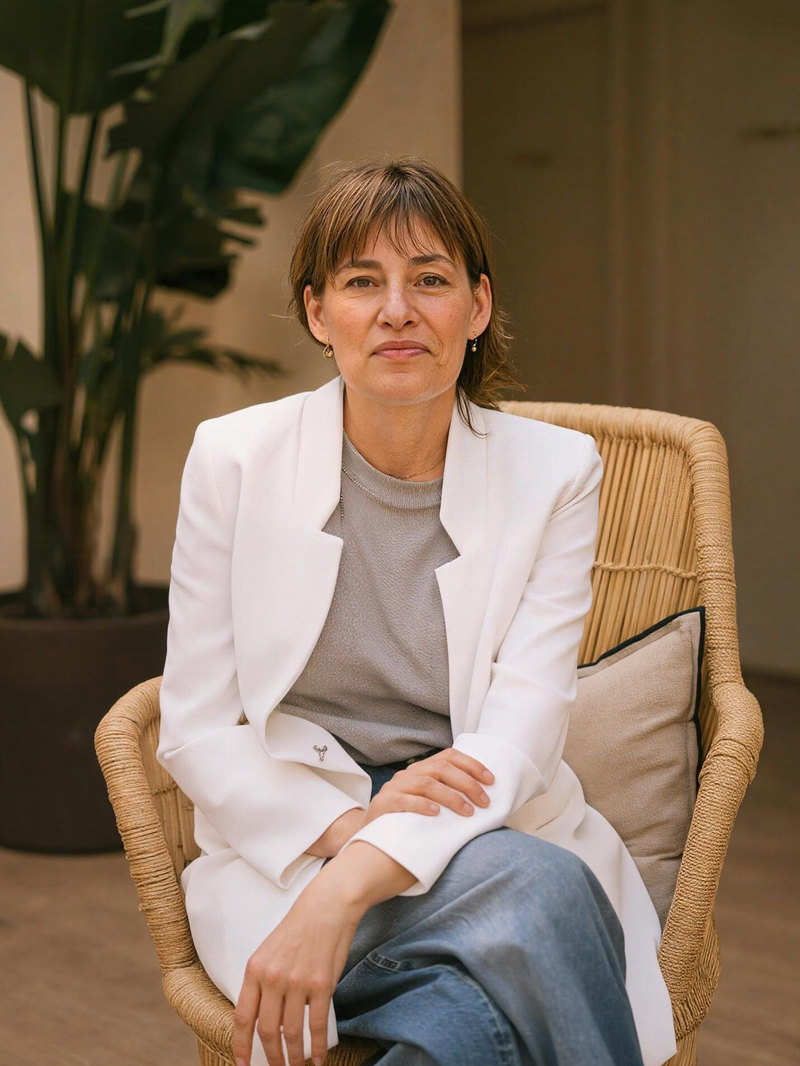 Montse Carreras, sentada en una silla de mimbre con americana blanca, mirando a cámara.