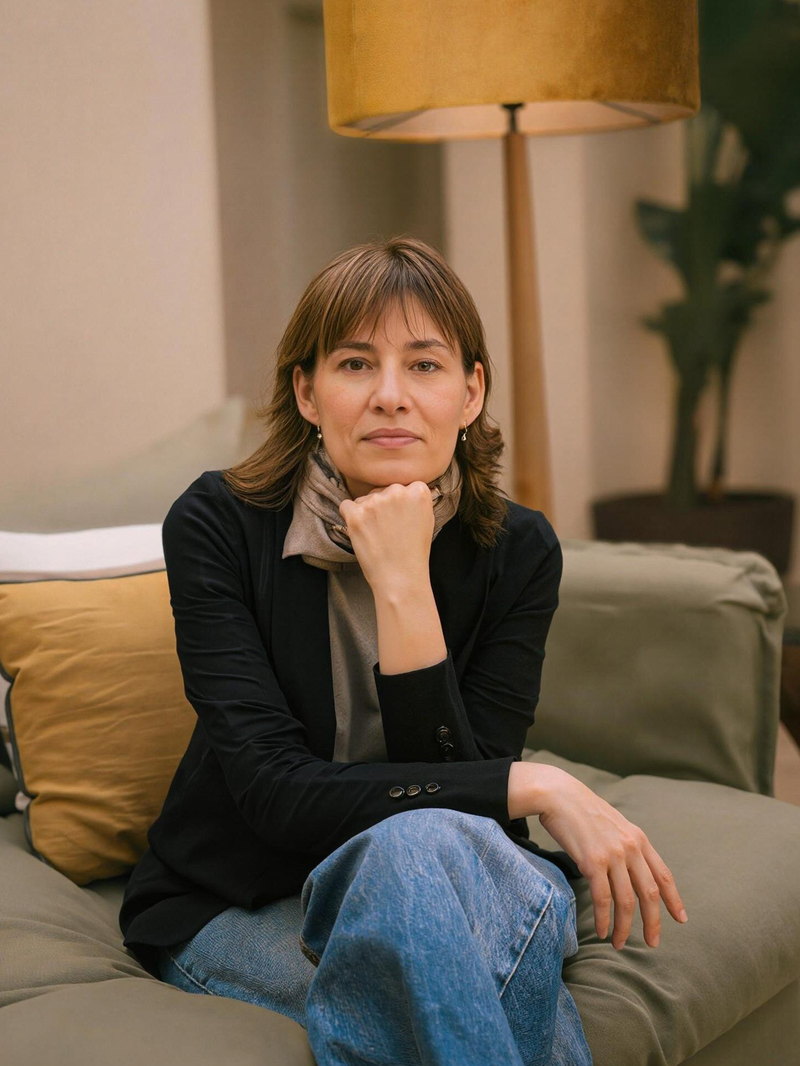Montse Carreras sentada en un sofá, con la mano bajo la barbilla, en una sala con luz cálida.